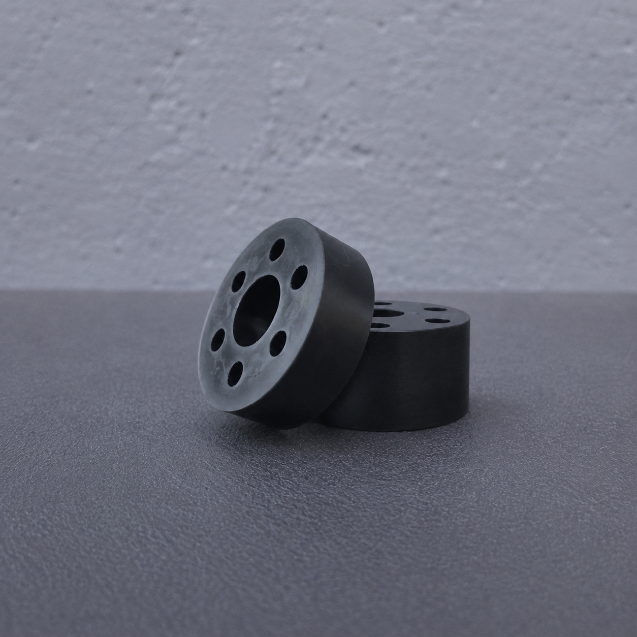 Two black circular cylindrical components with multiple circular cutouts, standing upright on a dark textured surface against a light gray wall.